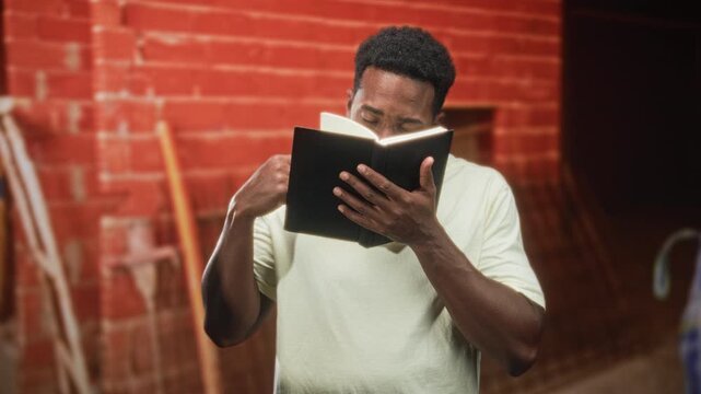 Man reads book and turns page with hands while standing against a red brick wall in a building; contemplation focus.