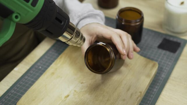 Close-up of female hands heating glass jars while making homemade wax candles in a workshop using a heat gun.