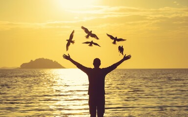 Fototapeta premium Man standing in water with arms outstretched as seagulls fly around him at sunset