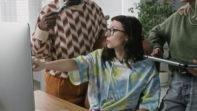 Young female team mentor pointing at computer screen guiding two senior colleagues through digital workflow while sitting at desk in modern office