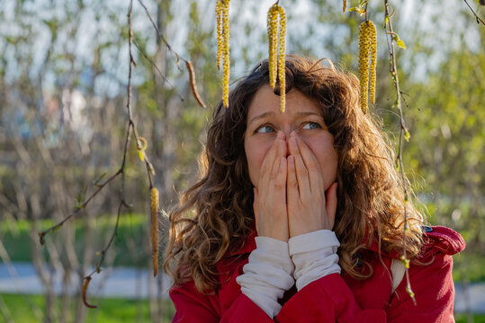 Young woman with curly hair wearing a red coat is covering her nose and mouth with her hands, suffering from seasonal pollen allergy caused by birch tree catkins during springtime in a park
