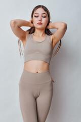Fototapeta premium Young woman in beige sportswear stretching with hands behind head, eyes closed, standing against neutral background for fitness and wellness. Relaxed, healthy lifestyle and calm pose.