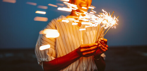 A woman stands under a dark blue sky, holding sparklers in her hands. The sparklers create bright sparks as she enjoys a celebration. This scene captures a moment of joy and excitement. © maxbelchenko