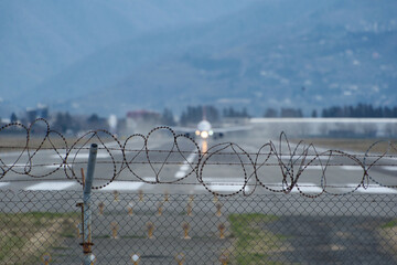 Barbed wire fence by the airport, blurry plane taking off and mountains beyond