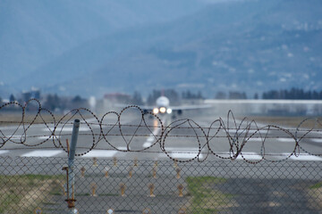 Barbed wire fence by the airport, blurry plane taking off and mountains beyond