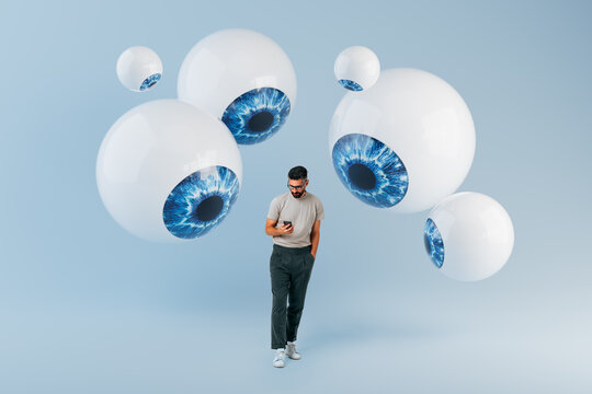 Man standing while using smartphone surrounded by floating 3D eyeballs in studio