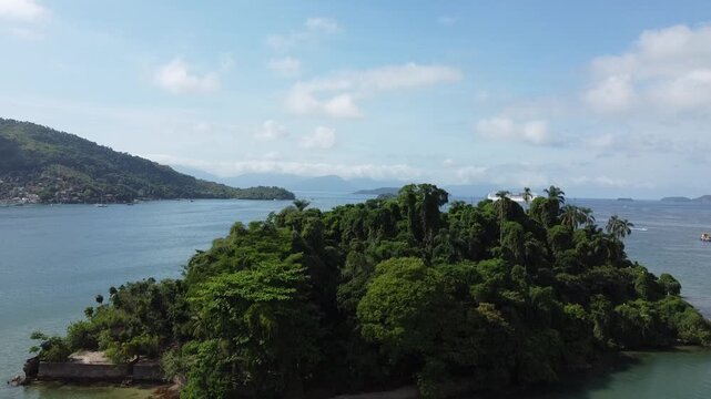 Drone rises behind the small tropical Island, covered in dense Atlantic rainforest, Ilha do Coqueiro in the Bay of Angra dos Reis, and reveal the stunning Angra dos Reis Bay behind it
