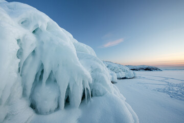 Natural ice formations and frozen splash patterns on a snowy winter landscape at sunset. Dramatic icicles and smooth snow surface create a minimalistic arctic scenery with soft pastel sky. Cold season © Olga Lyubochkina