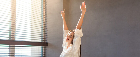 A woman stands beside a window with her arms raised. She enjoys the sunlight streaming in. The room...