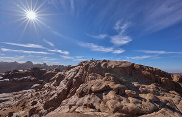 Obraz premium Mountain landscape with a female tourist posing in a summit in the national park Saint Catherine Protectorate, Sinai’s highlands. Red granite mountains. Vacation in Egypt. Rays radiating from the sun