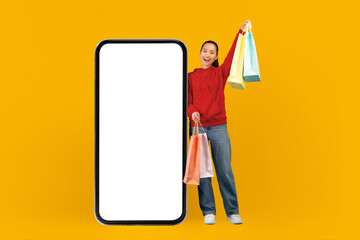 A woman stands beside a large smartphone screen holding several shopping bags. She appears happy...