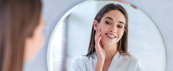 A woman stands in front of a mirror in a light room. She is smiling and touching her face. The reflection shows her interest in her appearance. © Prostock-studio