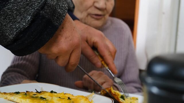 Man hands use plate and fork to feed an elderly grandmother with wrinkled but lively face. Elderly care, senior support, retirement home, and happy aging concept.