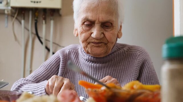 Very elderly woman with wrinkled face and gray hair calmly eats food at a table using fork and fingers. Elderly care, special homes for homeless seniors, and caring for aging parents.