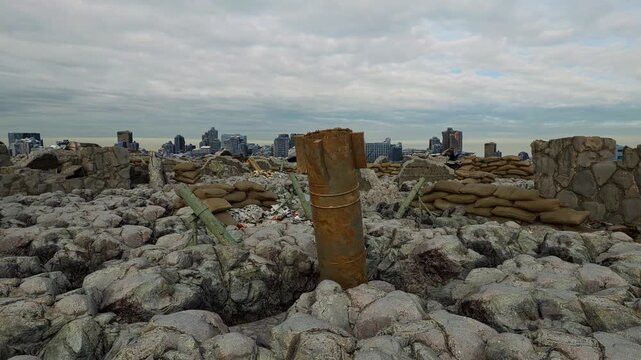 An unexploded aerial bomb stands upright in battlefield rubble, with a war-torn city skyline in the distance.