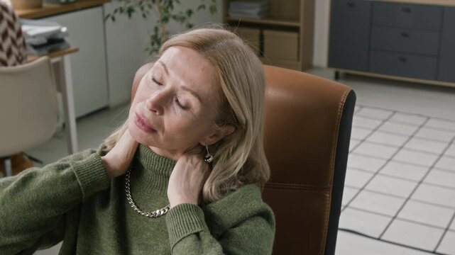 Overwhelmed senior female manager removing glasses then stretching stiffened neck muscles after hours of computer work in open space office