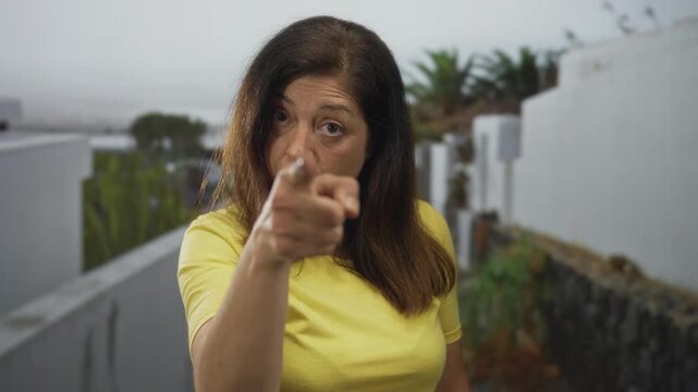 Woman in yellow shirt points index finger sharply along a white building wall outdoors on a balcony walkway to guide focus; command.