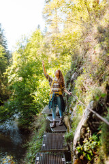 Fototapeta premium A female hiker with a backpack walks along a path in a rocky forest. A young woman on a hike, enjoying the sunset and the adventure in the mountains. Active lifestyle. Concept: recreation, hiking.