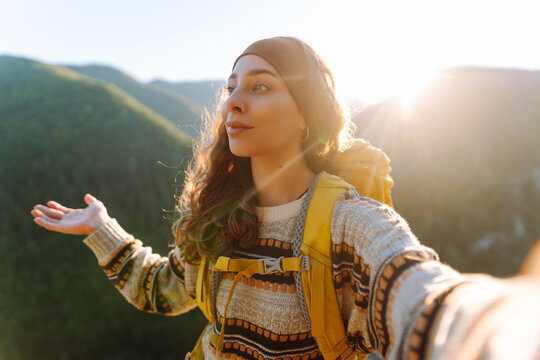 A cute woman with a hiking backpack takes a selfie on the edge of a cliff on a sunny day. A beautiful woman vlogs while traveling and enjoys the sunset. Concept: technology, travel.