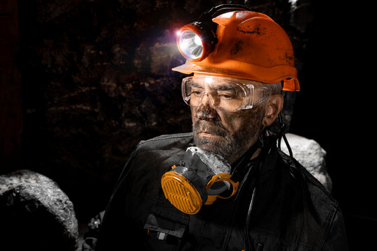 Coal miner with a dirty face in black dust, wearing a protective helmet with a flashlight, filtering respirator and work clothes for working in mine. Hard work underground in a coal mine, digging site