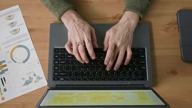 Top view of hands of senior woman entering data on laptop while managing paperwork at desk in office workspace