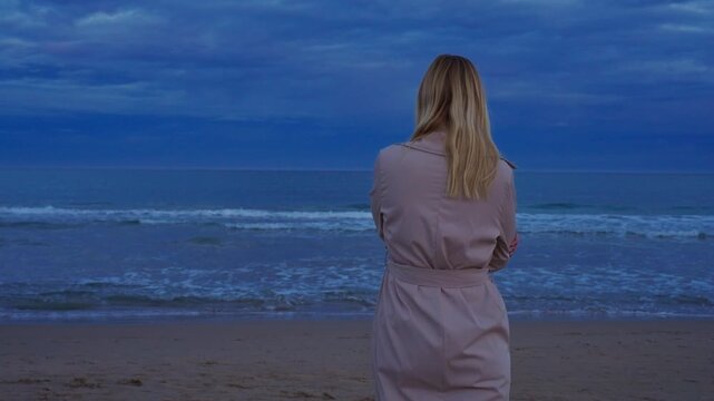 Back view of lonely blonde woman in trench coat stand on sandy beach watch dark blue sea. Solitude and contemplation during cold, cloudy evening.Concept of choice, uncertainty,reflection and important