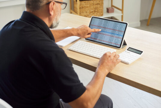 Senior man accountant reviews spreadsheet on tablet. At a desk, he analyzes finance and business accounting with a calculator at home office. Remote money management and budgeting concept.