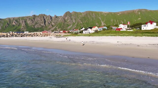 forward drone flight over sea towards sandy beach on Vesteralen, Vester&aring;len islands. Caucasian man walking with bare feet on beach. Tourist enjoying summer in Norway. Bleik village en mountains in bac