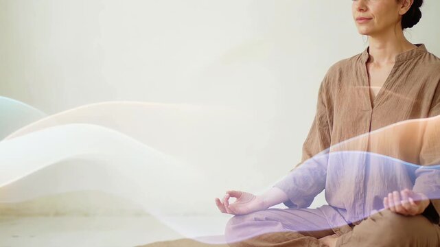 Asian woman in beige meditation attire sits cross-legged on a mat, hands raised in a meditative gesture, with flowing light patterns surrounding her in a serene indoor setting