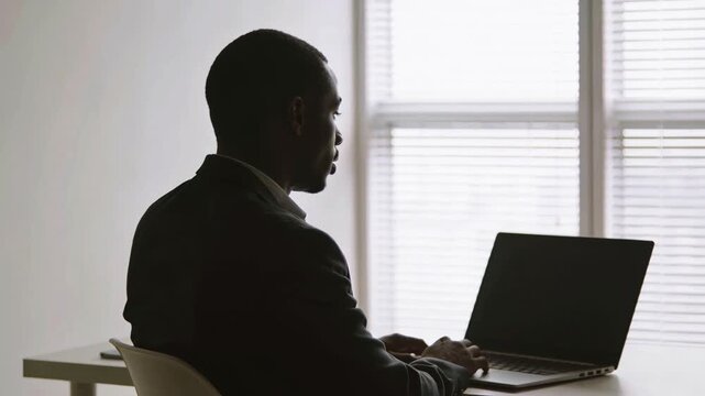 African American man in a suit sits at a desk working on a laptop, with sunlight filtering through blinds in a modern office space