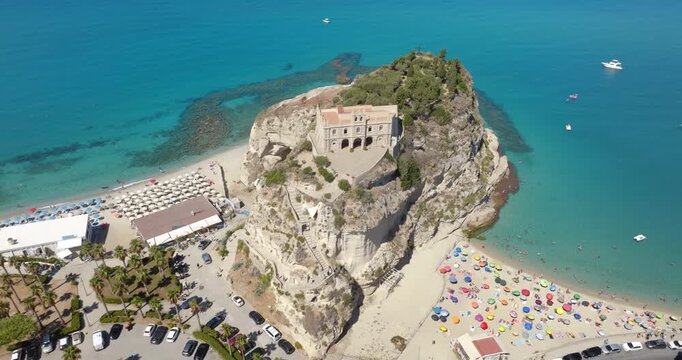 Aerial view of Sanctuary of Santa Maria dell'Isola in Tropea, Calabria, Italy. The ancient church stands on a rocky promontory, overlooking a crowded sandy beach filled with colorful sun umbrellas.
