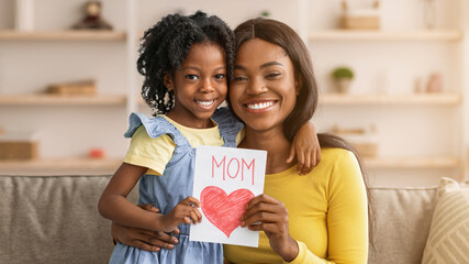 A young girl stands next to her mother, holding a drawing with the word mom and a heart. They both smile joyfully in a warm living room. The scene shows family love and connection. © Prostock-studio