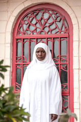 A woman in a white hijab stands solemnly before a red arched doorway