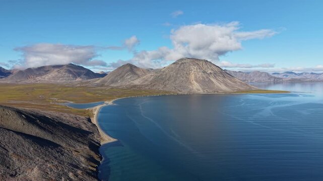 Aerial drone footage reveals side tracking along a curved sandspit at Rumilet Bay. The camera holds wide water and tundra in Chukotka during early autumn. Sunlit clouds add scale and depth
