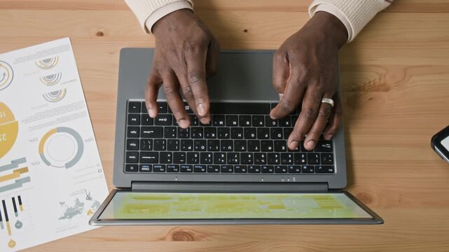 Top view of hands of senior Black man entering data on laptop while working with documents at wooden desk in modern office