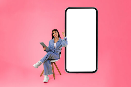 Full boody length vertical portrait of smiling young woman using tablet sitting on chair isolated on orange studio background, looking at camera and pointing finger up, showing copy space