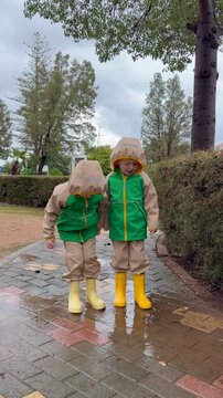 Two boys in raincoats and rubber boots jump in puddle on park path, kids activity footage for family lifestyle promotion, education projects and social media content about outdoor childhood play