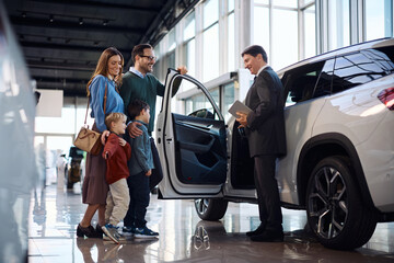Naklejka premium Happy car salesman talking to kids and their parents while showing them new family vehicle in showroom.