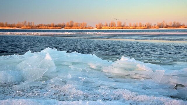 Stunning natural landscape showing ice floes drifting along the Dnieper river during the spring thaw, with the far bank illuminated by the warm light of the setting sun and birds flying overhead