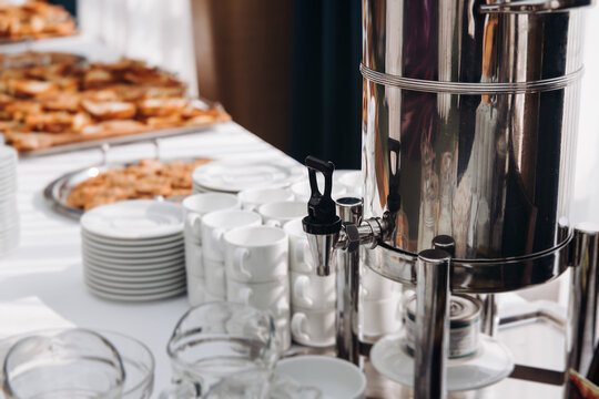 Set of coffee break lunch in the hotel restaurant during conference meeting, with tea and coffee catering, decorated banquet table with white cups, tableware and metal tea maker machine boiler