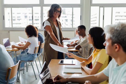 Teacher distributing exams while students sit at desks. Class receiving written academic tests in a high school classroom. Education and evaluation concept.