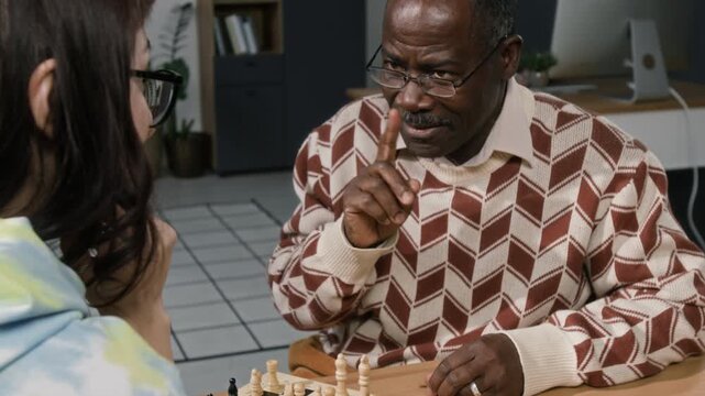 Focused senior African American man moving chess piece playing intellectual board game with young female colleague while having fun at workplace