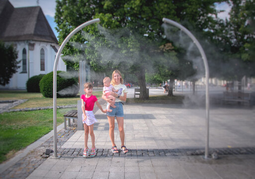 Happy family with two children cooling down near water misting station on a hot summer day. Mother holding infant while older daughter plays with refreshing spray. Family lifestyle and summer vacation