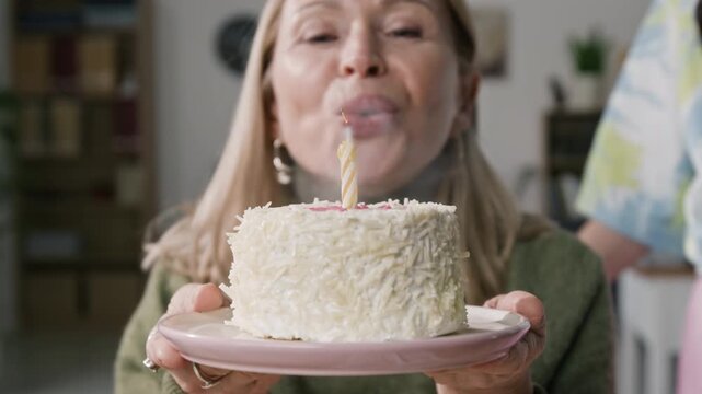 Close up on birthday cake in hands of senior woman blowing candle and making wish while celebrating occasion with colleagues at workplace, slow motion