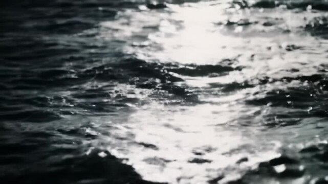 Extreme close-up overhead video of dark, abyssal ocean water surface, dynamically churning with agitated waves, glistening reflections and shimmering streaks in low-key light, deep ocean mystery