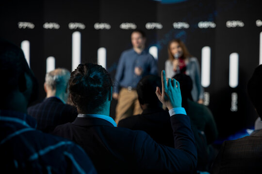 Male listener in audience asking questions during presentation and panel discussion, public speakers team engaging in a debate with attendees. Raising hand and being active in conference hall.