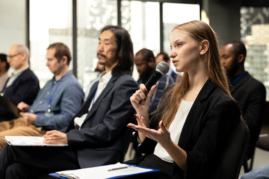 Young woman journalist reading questions from clipboard and taking notes, interacting with presenter via microphone in conference room. Writing answers for press purposes at business summit.