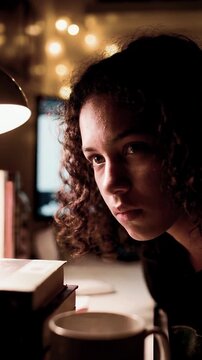 Studious young woman with curly hair looking at computer screen in dark room with warm desk lamp for online education distance learning and academic research concept