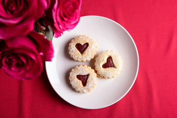 Over head view and Close-up of a heart-shaped jam-filled cookie dusted with powdered sugar and served on a white plate. The soft focus background and warm red tones create a romantic, cozy mood, makin