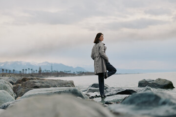 A woman in a grey coat stands on rocks by the sea, looking thoughtfully at the horizon under a cloudy sky. Coastal landscape with mountains and calm water. © SHOTPRIME STUDIO